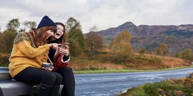 Two ladies sat on car bonnet looking and taking photos of the mountains