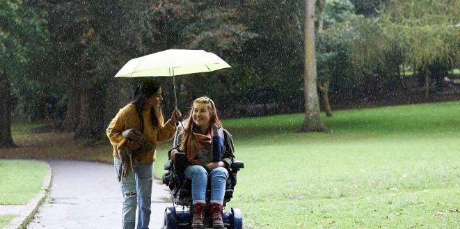 Two ladies on a walk one lady in a mobility scooter and other lady holding umbrella over them both